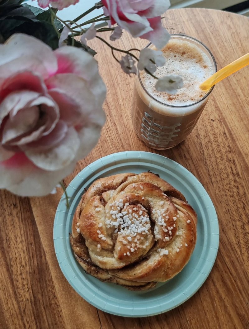 Voisilmäpulla lautasella sekä maitovaahdolla koristeltu kahvi puisella pöydällä, kukkia taustalla.