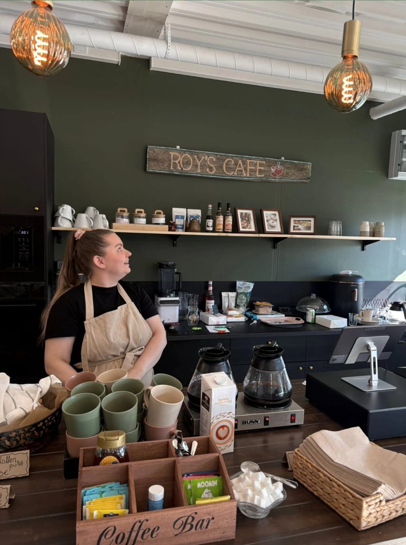 Roy’s Cafén yrittäjä kahvilan tiskin takana, taustalla vihreä seinä, messinkiset lamput ja Roy’s Cafe -kyltti.